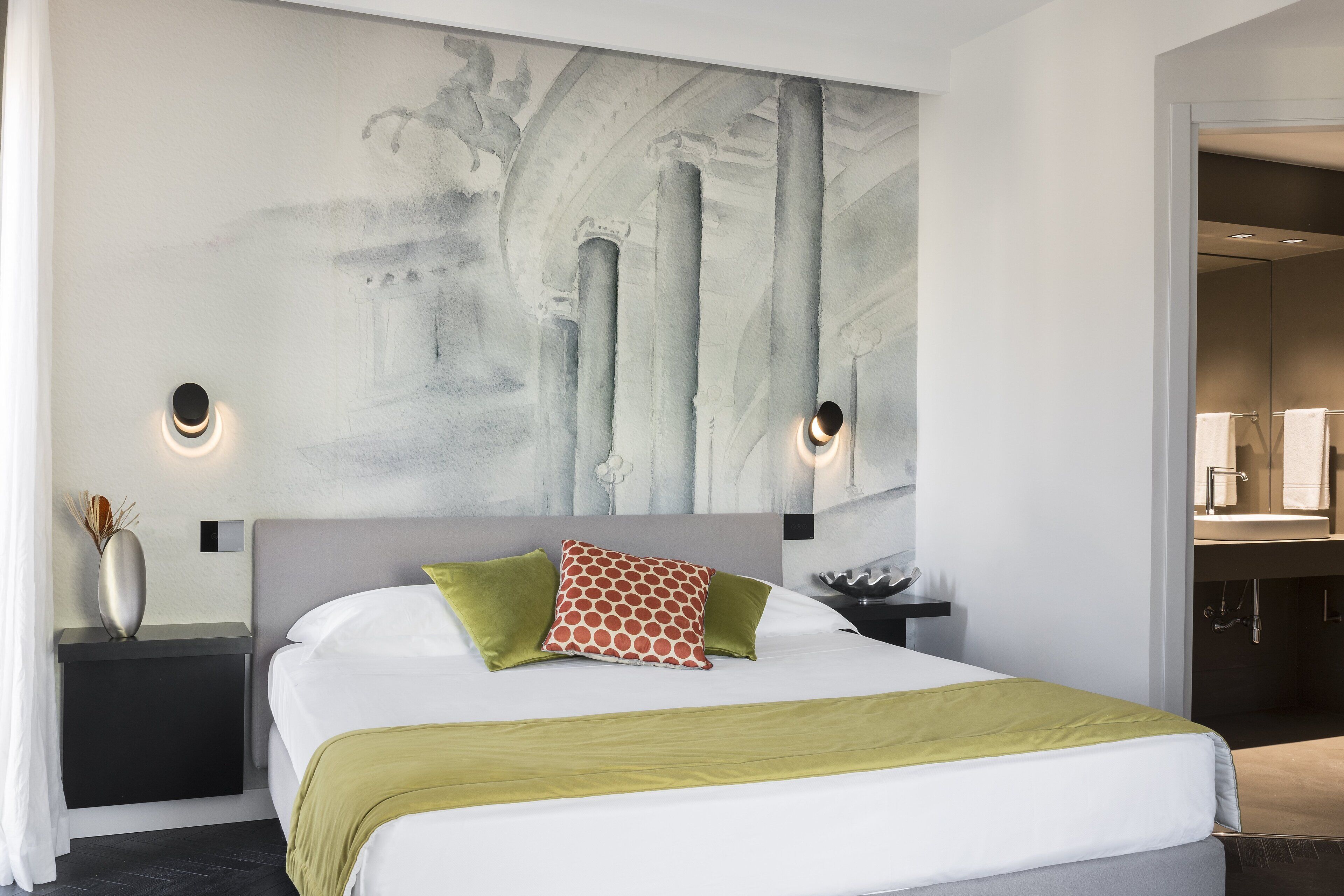 Et moderne hotelværelse med en stor seng, et natbord, en vægmonteret lampe og et badeværelse, der kan ses igennem en åben dør.