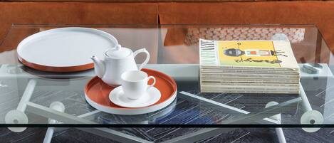 A glass-top coffee table with a white teapot and saucer, a tray, and a stack of magazines.