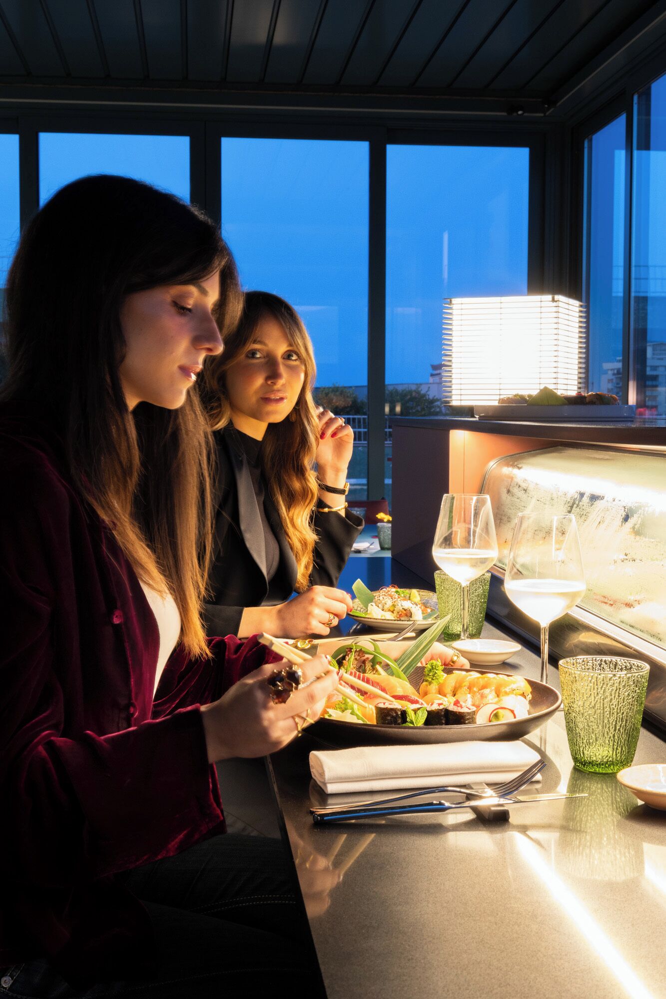 Two people dining at a table with a city view, a glass of white wine, and a plate of sushi.