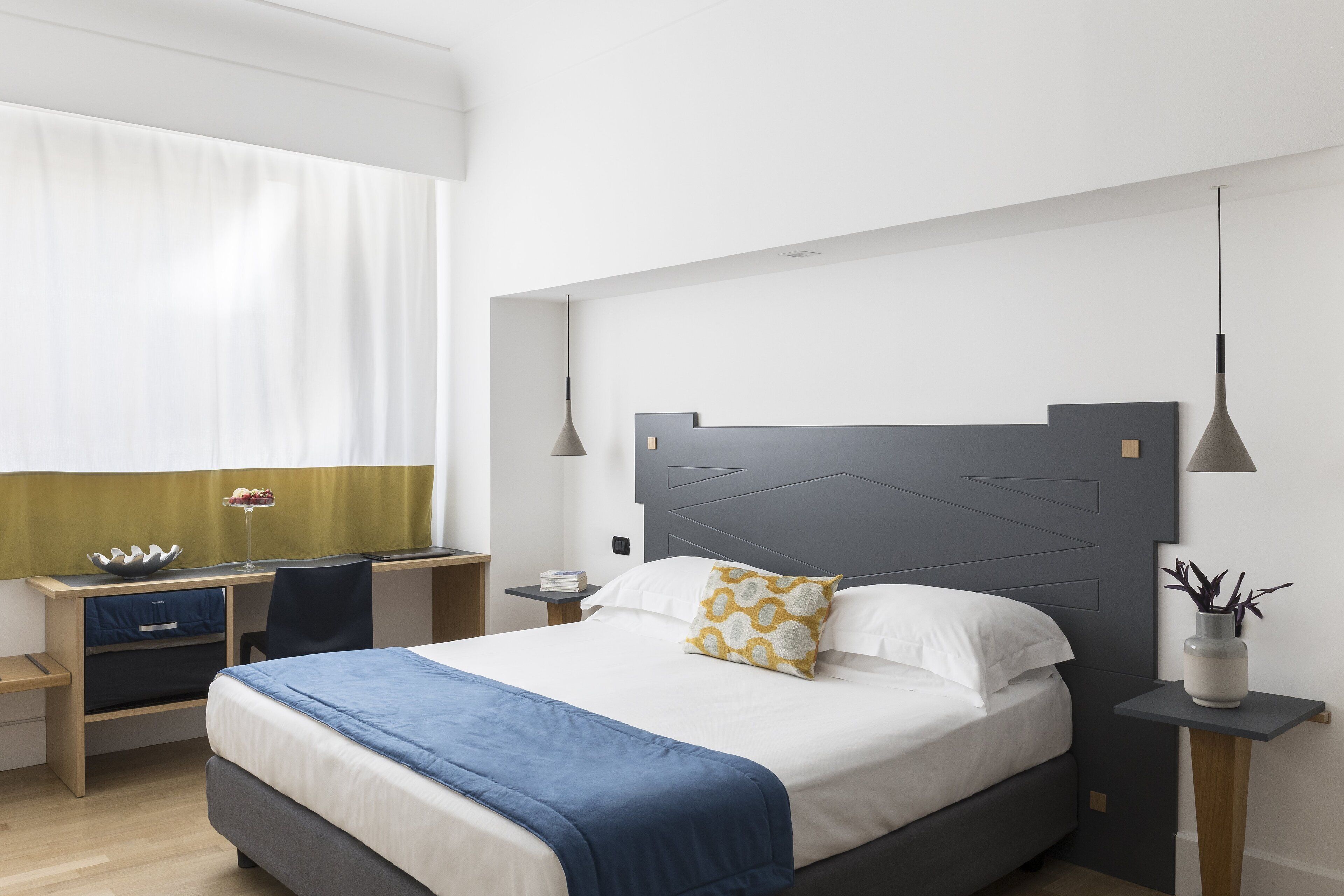 A modern hotel room with a large bed, a desk, and a chair. There is a window with curtains, a bedside table with a lamp, and a plant on a side table.