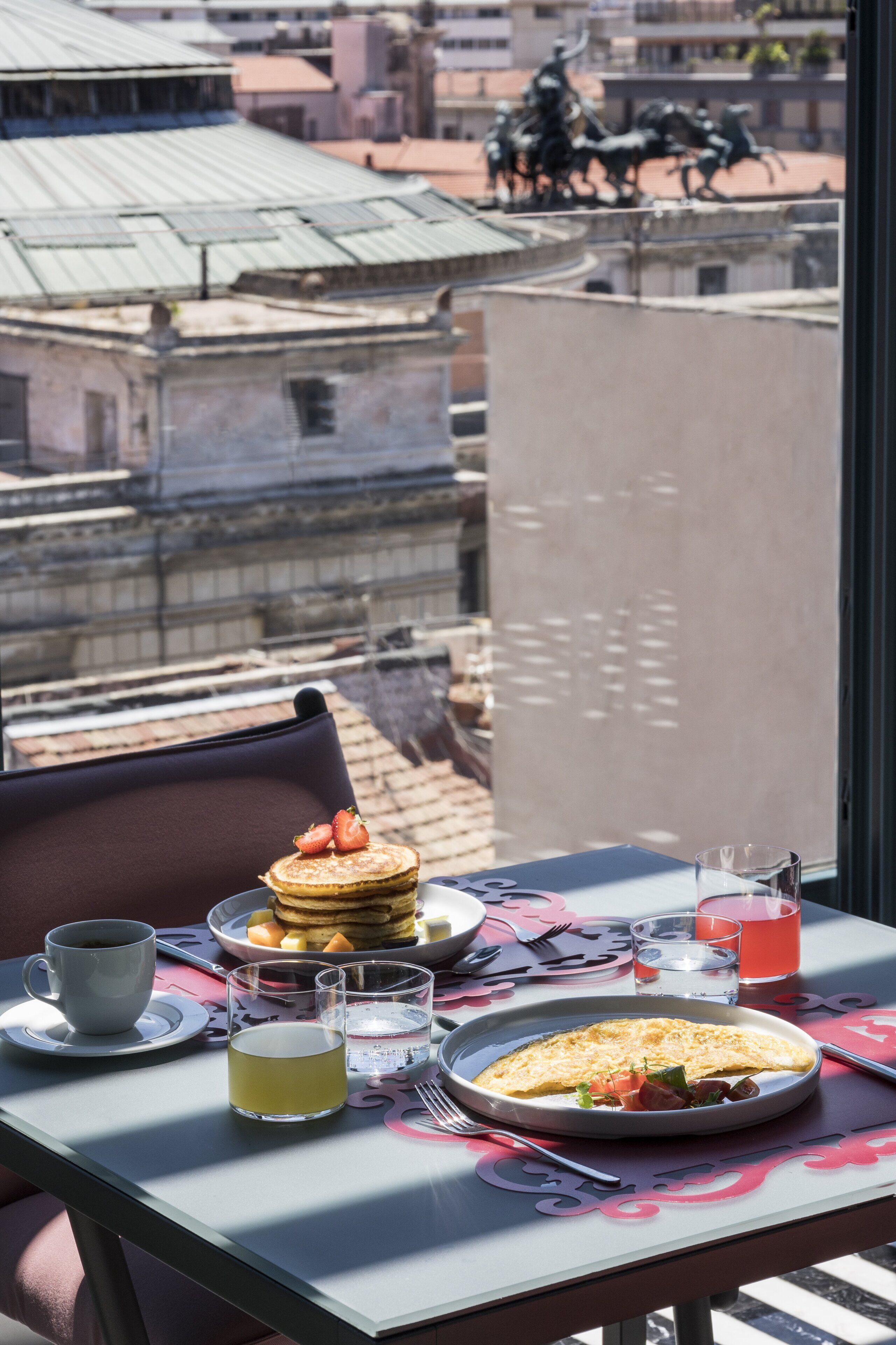 Mesa de desayuno con panqueques, un plato de comida y bebidas, con vista a un paisaje urbano que incluye edificios y una estatua.