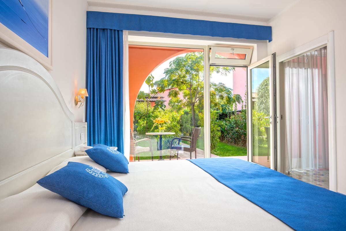 A bedroom with a bed, blue pillows, a nightstand, and a window with a view of a garden and a building.