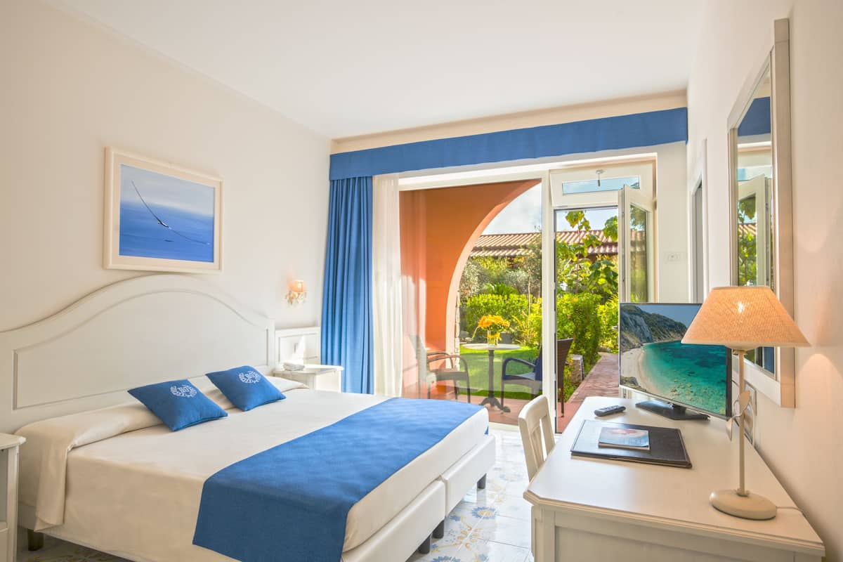A hotel room with a bed, a desk with a lamp, a TV, and a view of a garden through a glass door.