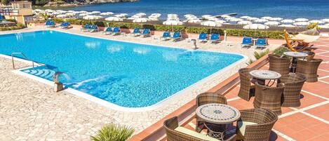 Un área de piscina con sillones de descanso y sombrillas, vista despejada del mar y un patio empedrado.