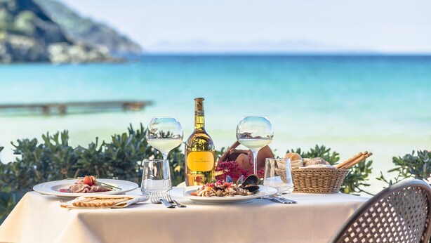 Mesa lista para una comida con una botella de vino, platos de comida y vasos, con vista a una playa de aguas cristalinas y un litoral rocoso.