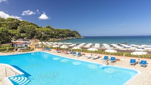 Una piscina con sillones de descanso y sombrillas, una playa y montañas al fondo.