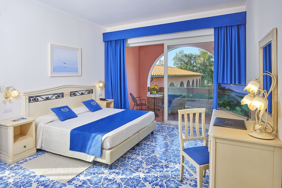 A hotel room with a bed, a desk, a chair, a nightstand, and a view of the outside.