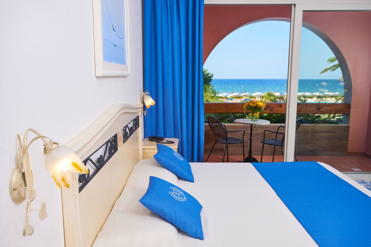 A hotel room with a bed, a nightstand, a lamp, and a view of the ocean through an arched window.