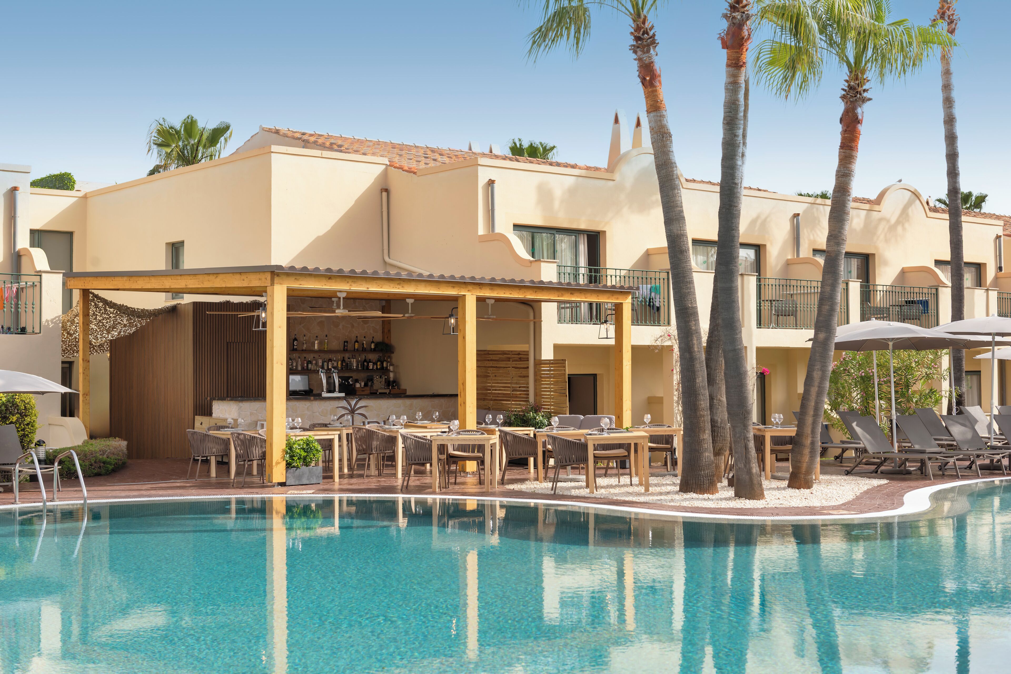 A pool area with lounge chairs and a bar, surrounded by palm trees and a building with a tiled roof.