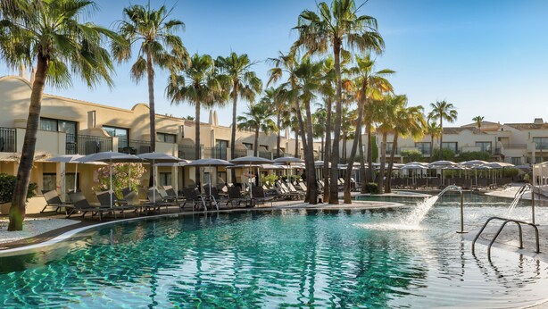 Una piscina con sillas de descanso y sombrillas, rodeada de palmeras y edificios de hotel.