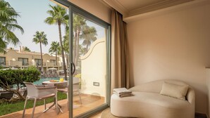 Una habitación de hotel moderna con un sofá curvo, un ventanal de vidrio que da a una zona de piscina y vistas a palmeras y otros edificios.