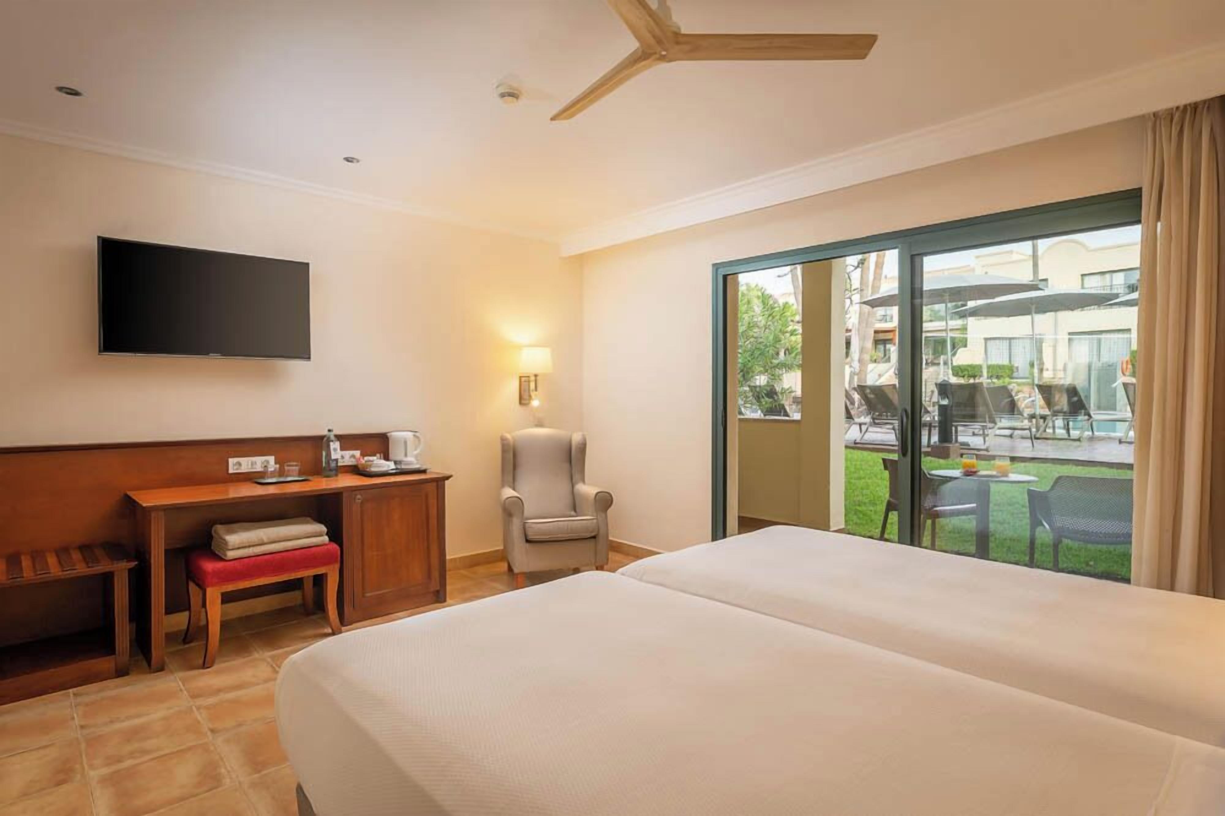 Ein Hotelzimmer mit einem großen Bett, einem Schreibtisch mit Stuhl, einem Fernseher und Blick auf eine Außenpatio mit Tischen und Stühlen.