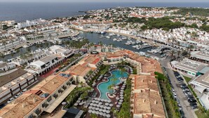 Un resort costero con un puerto deportivo, piscina y varios edificios de arquitectura mediterránea.