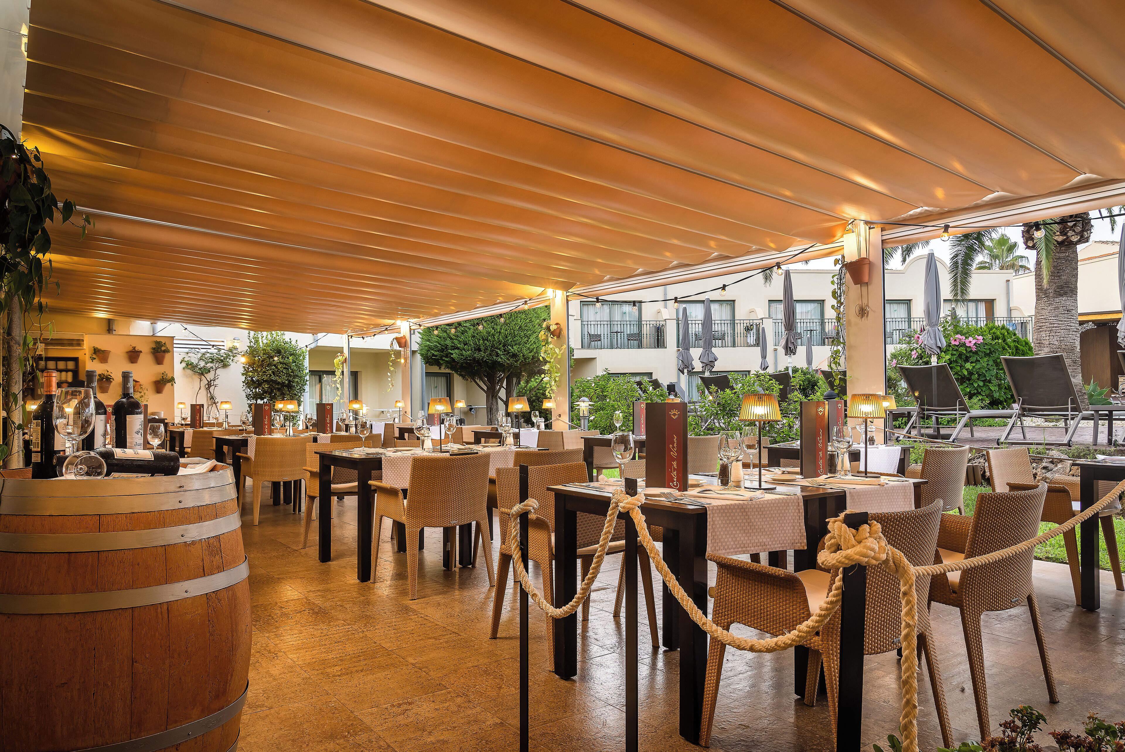 Un restaurante al aire libre con mesas, sillas, un barril de madera y plantas en macetas.