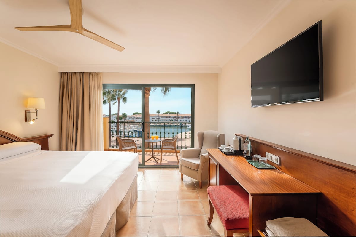 A hotel room with a bed, a desk, a chair, a TV, and a balcony with a view of palm trees and buildings.