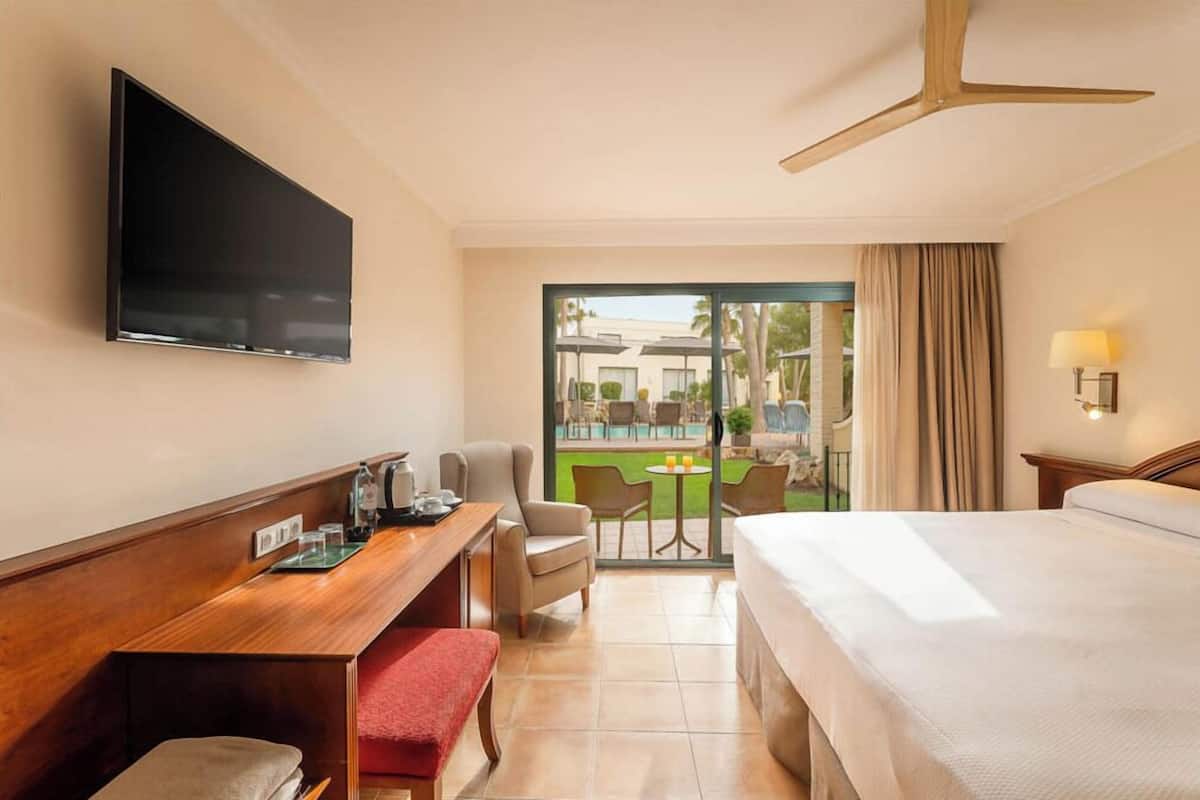 A hotel room with a bed, a desk with a flat-screen TV, a chair, a small table with a lamp, and a view of a pool and outdoor seating area.