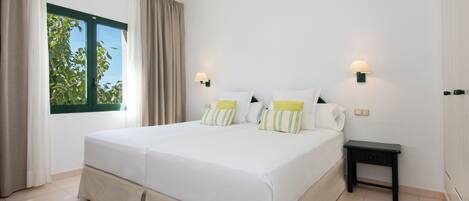 A double bed with white bedding and striped pillows, a bedside table with a lamp, and a window with curtains.