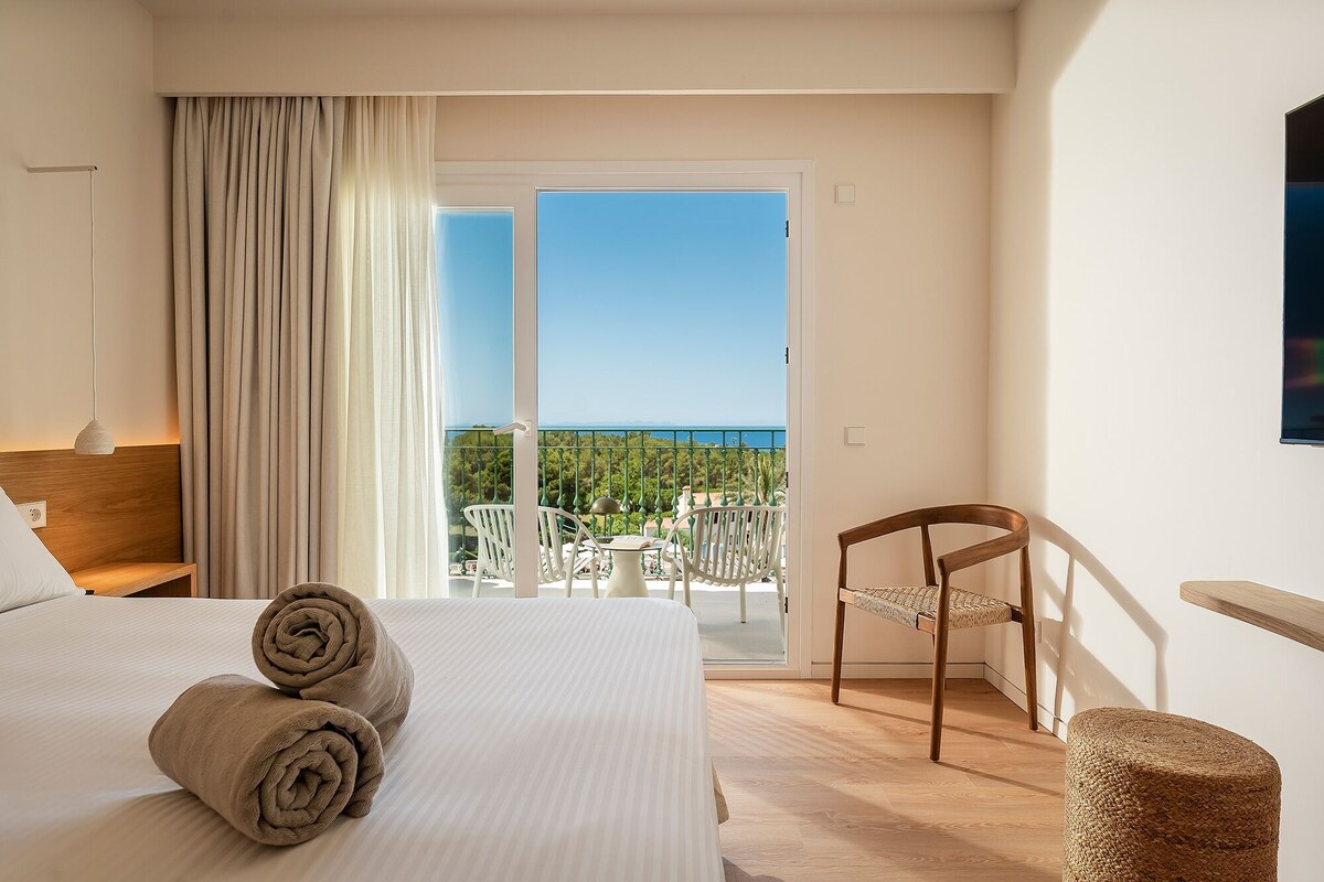 A hotel room with a bed, a chair, a small table, and a balcony with a view.