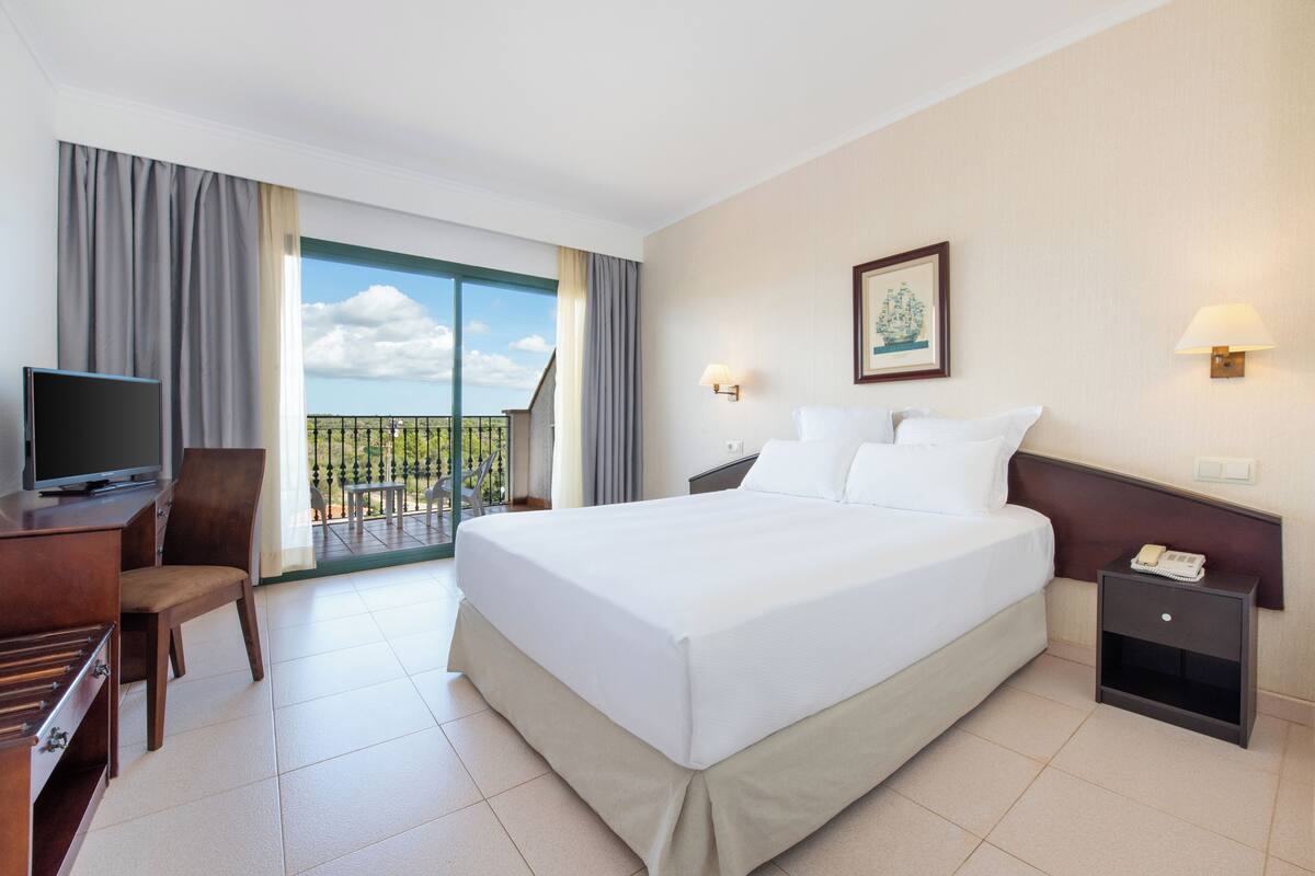 A hotel room with a large bed, a desk with a TV, a chair, a nightstand, and a balcony with a view.