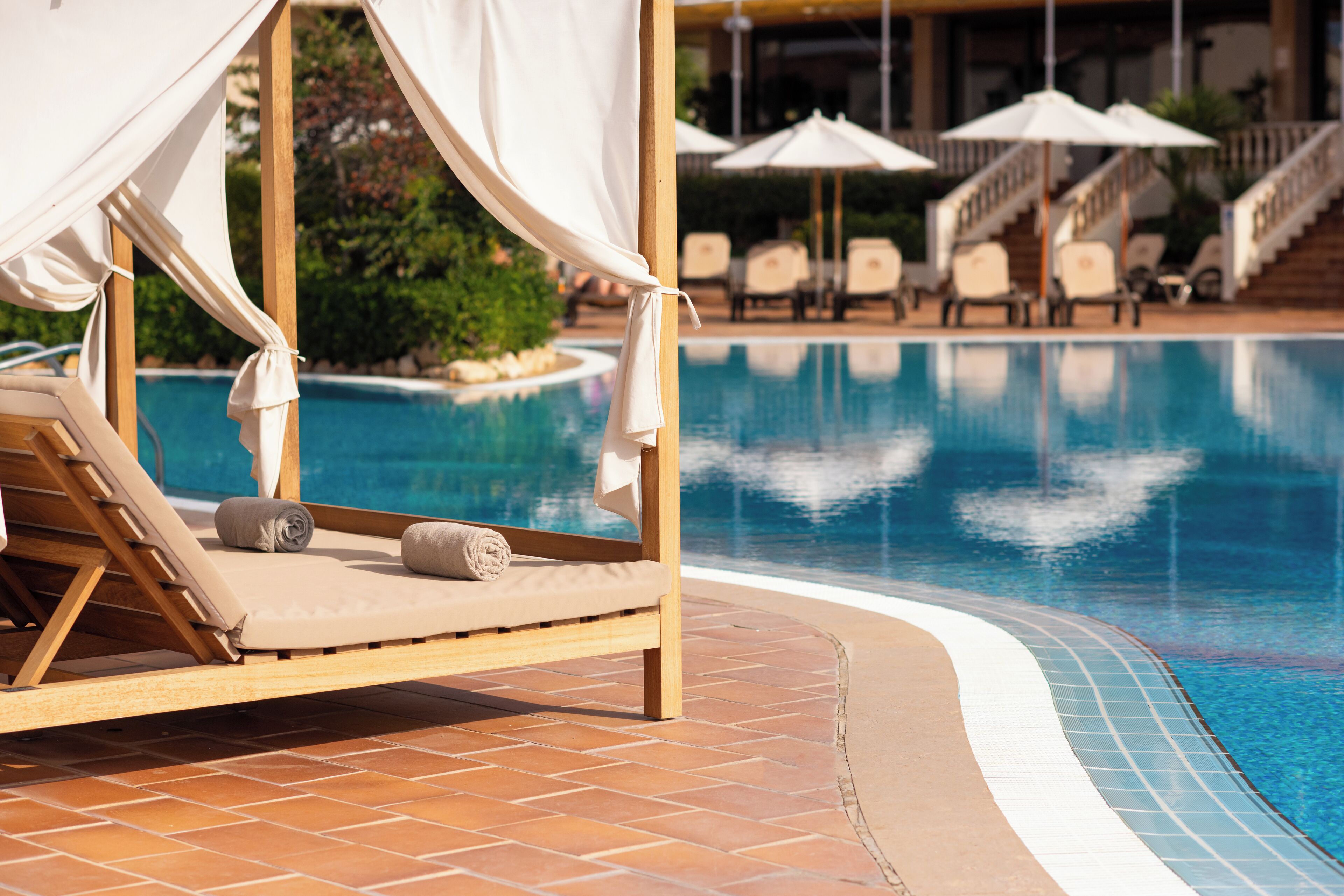 Ein Liegebereich am Pool mit einem hölzernen Baldachinbett, einer gefliesten Poolkante und Liegestühlen im Hintergrund.