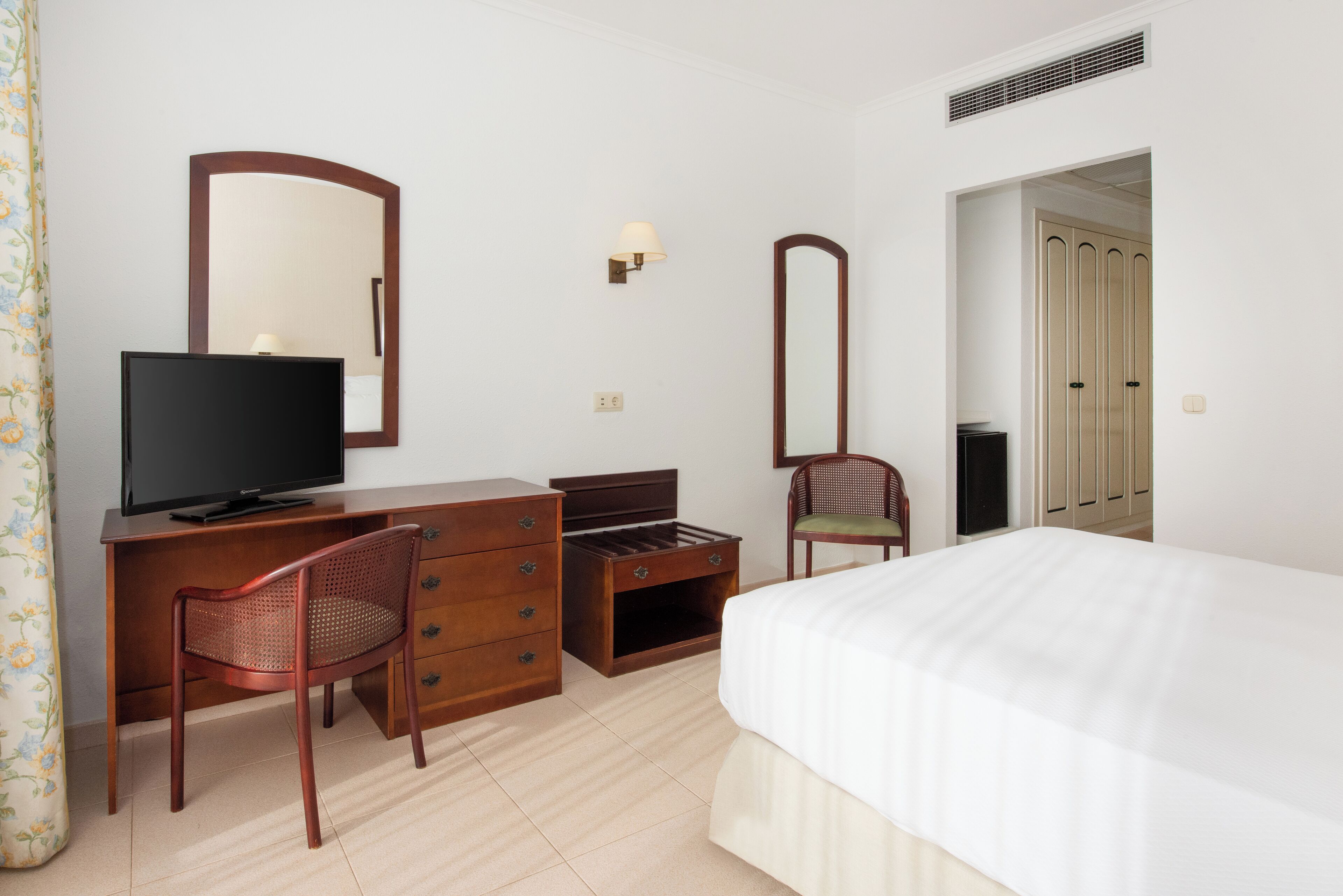 A hotel room with a bed, a desk with a television, a chair, a dresser, and a mirror.