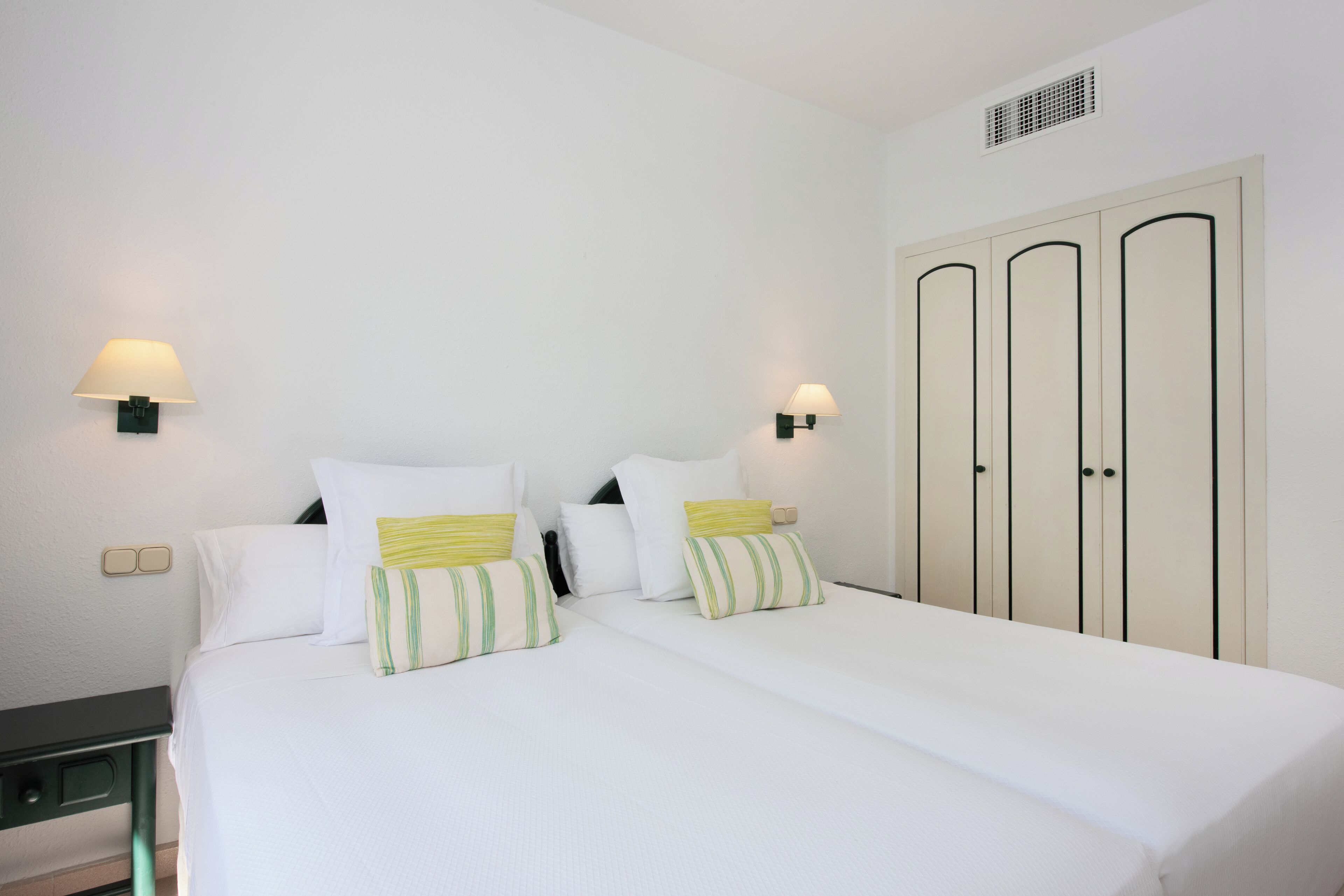Una habitación de hotel con una cama, dos lámparas de noche, un armario y una mesita.