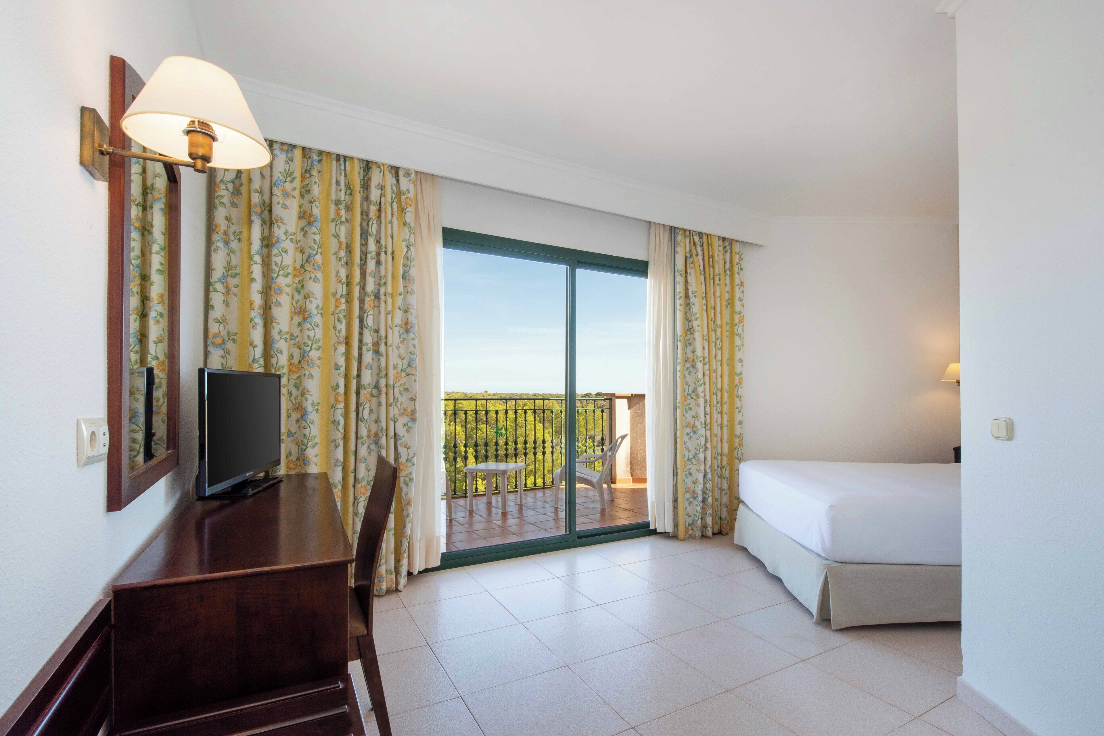 Ein Hotelzimmer mit Bett, Schreibtisch mit Fernseher, Balkon mit Tisch und Stühlen sowie Vorhängen.