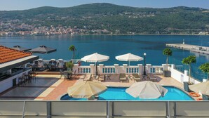 A rooftop pool area with lounge chairs and umbrellas, overlooking a body of water and a distant cityscape. - Samos Hotel  (Samos)