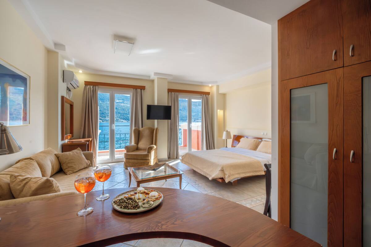 A hotel room with a bed, a sofa, a dining table with drinks, a TV, and a balcony with curtains.