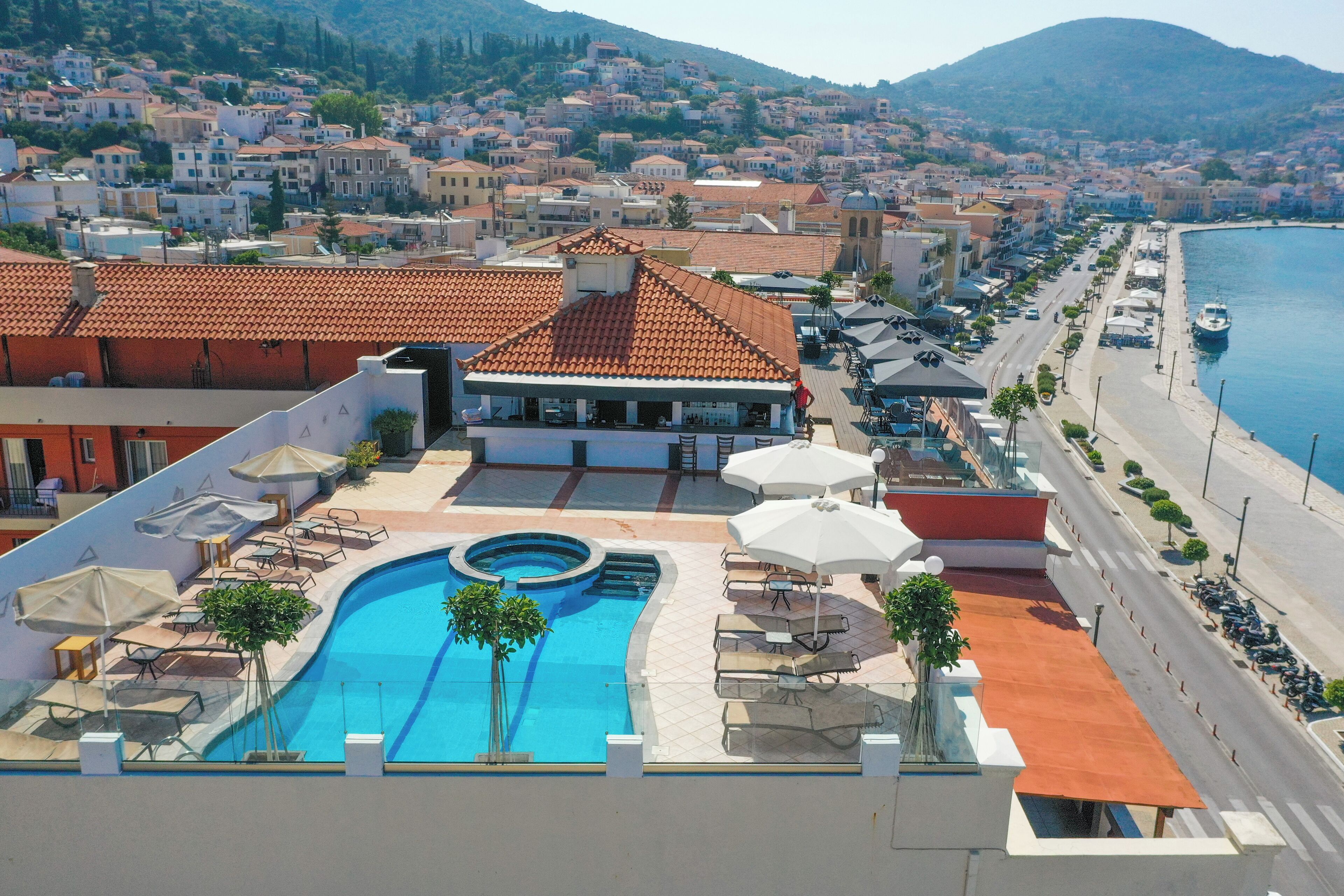 A rooftop pool area with lounge chairs and umbrellas, a swimming pool, and a view of a coastal town with buildings and a mountain in the background.
