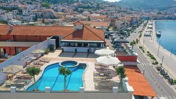 A rooftop pool area with lounge chairs and umbrellas, a swimming pool, and a view of a coastal town with buildings and a mountain in the background.