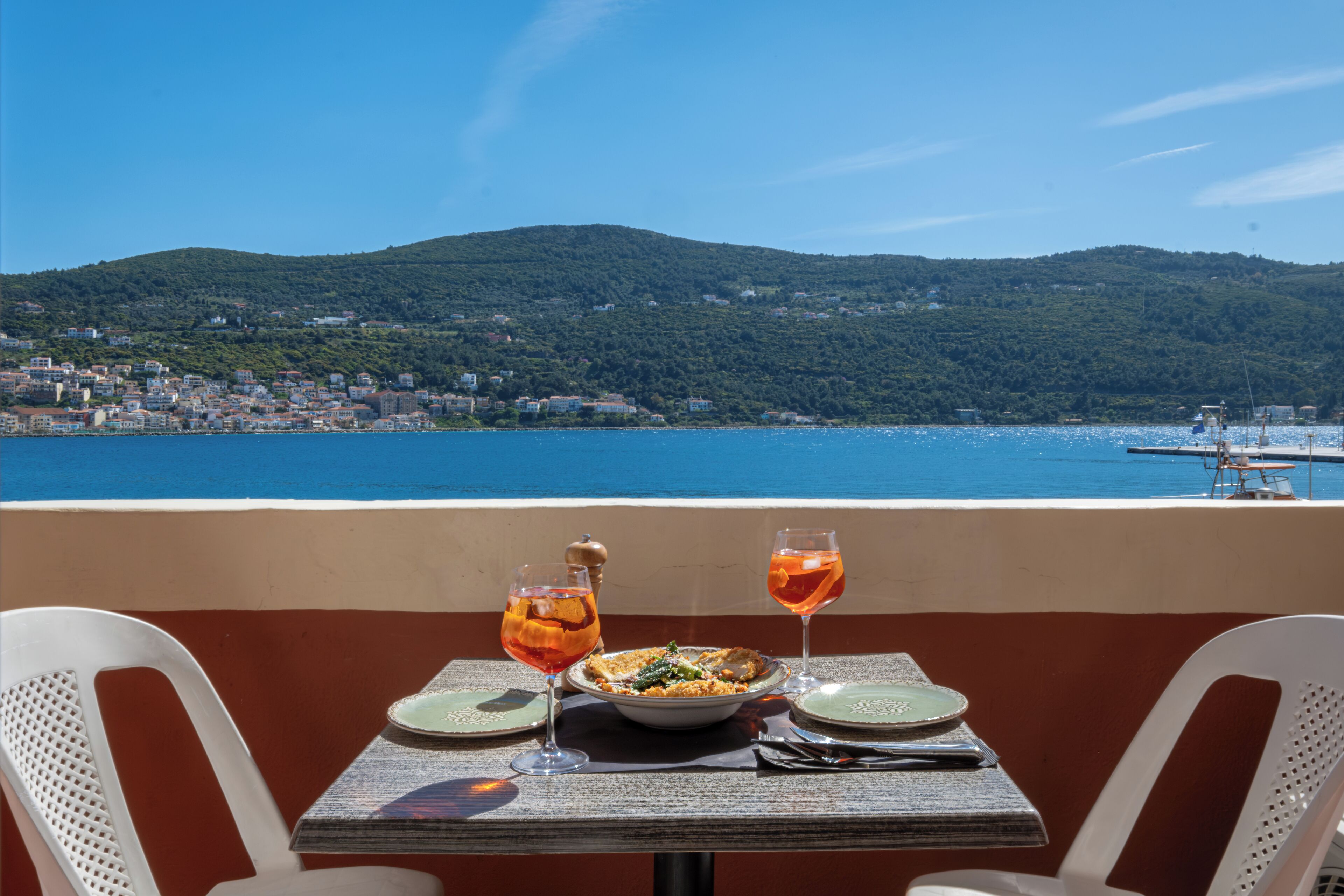 A table with a meal and drinks, overlooking a coastal town and hills.
