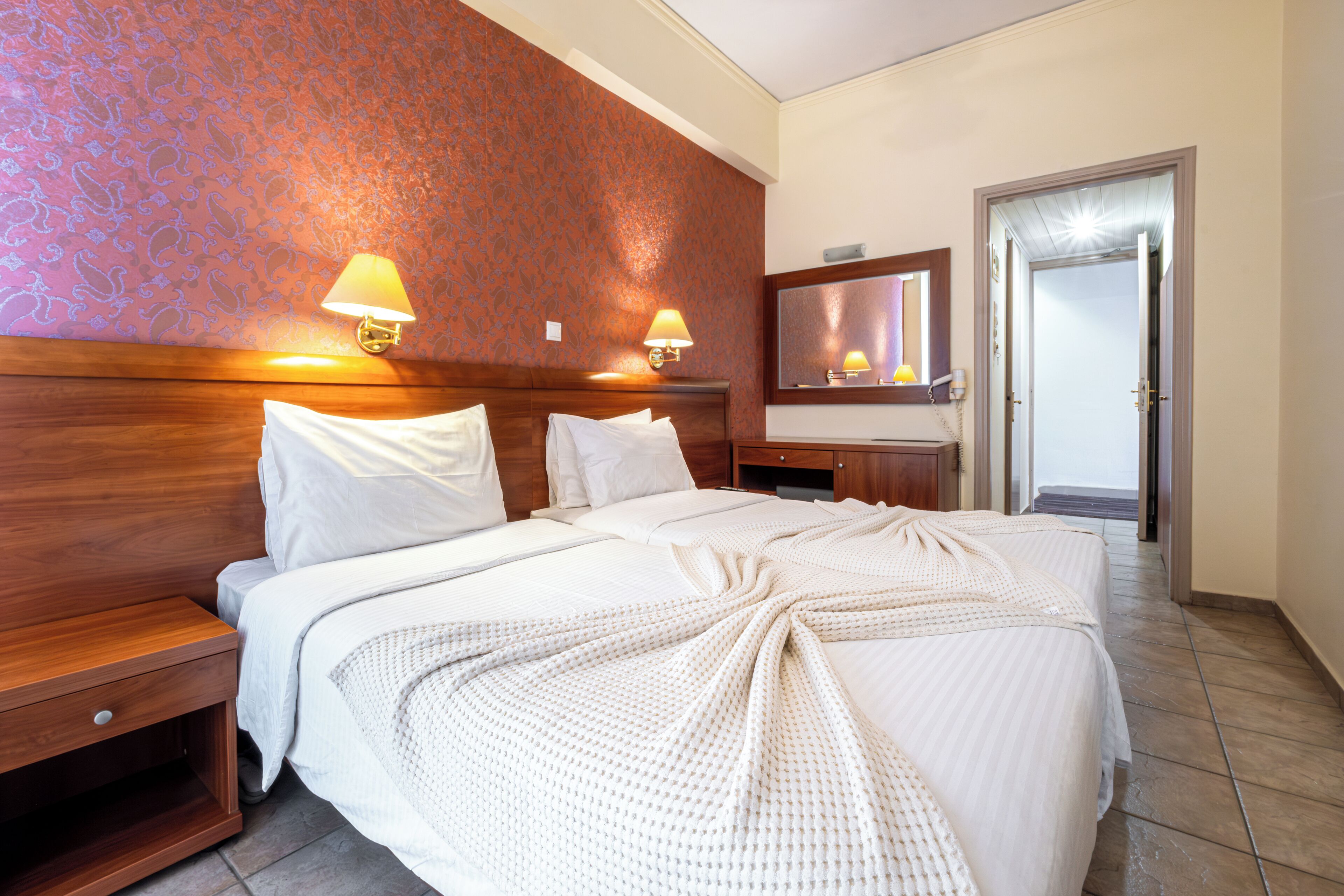 A hotel room with a large bed, two bedside lamps, a wooden headboard, a nightstand, a mirror, and a door leading to another room.