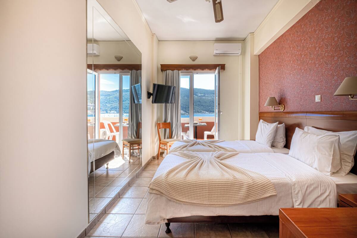 A hotel room with a large bed, a TV, a balcony with a view of the mountains, and a dining area with chairs.