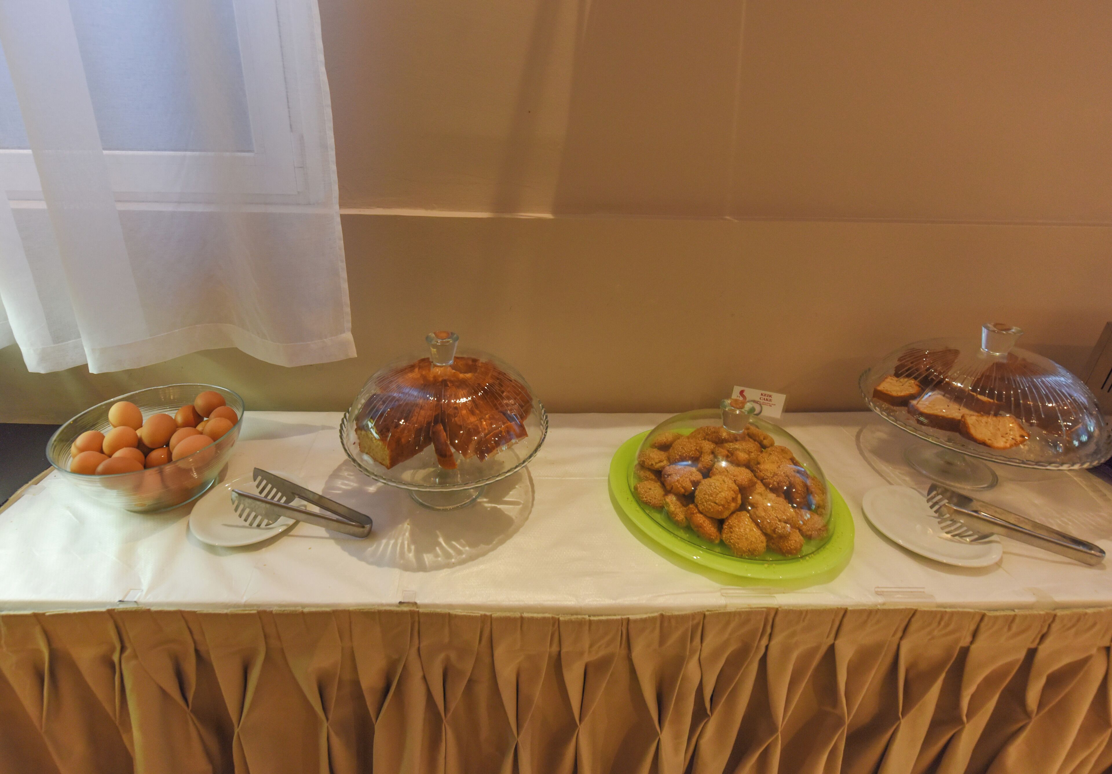 Una mesa de buffet con diferentes alimentos, incluyendo un tazón de huevos, un recipiente de vidrio con pasteles, y un plato verde con galletas.