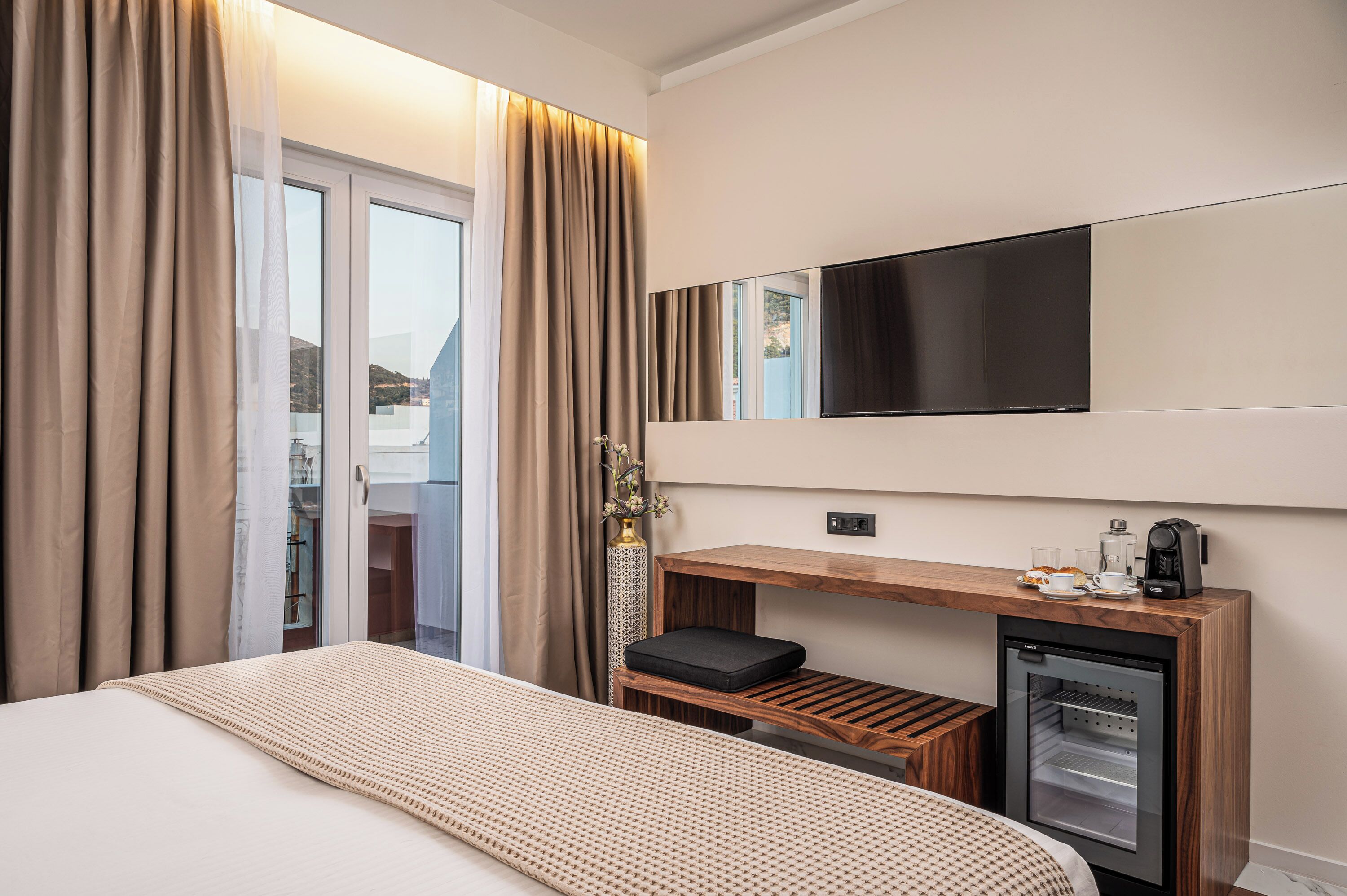 A hotel room with a bed, a small desk with a coffee maker, a mini-fridge, and a large window with curtains.