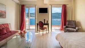 A hotel room with a bed, a sofa, a chair, a table with a vase, a TV, and a balcony with a view of the sea. - Samos Hotel  (Samos)
