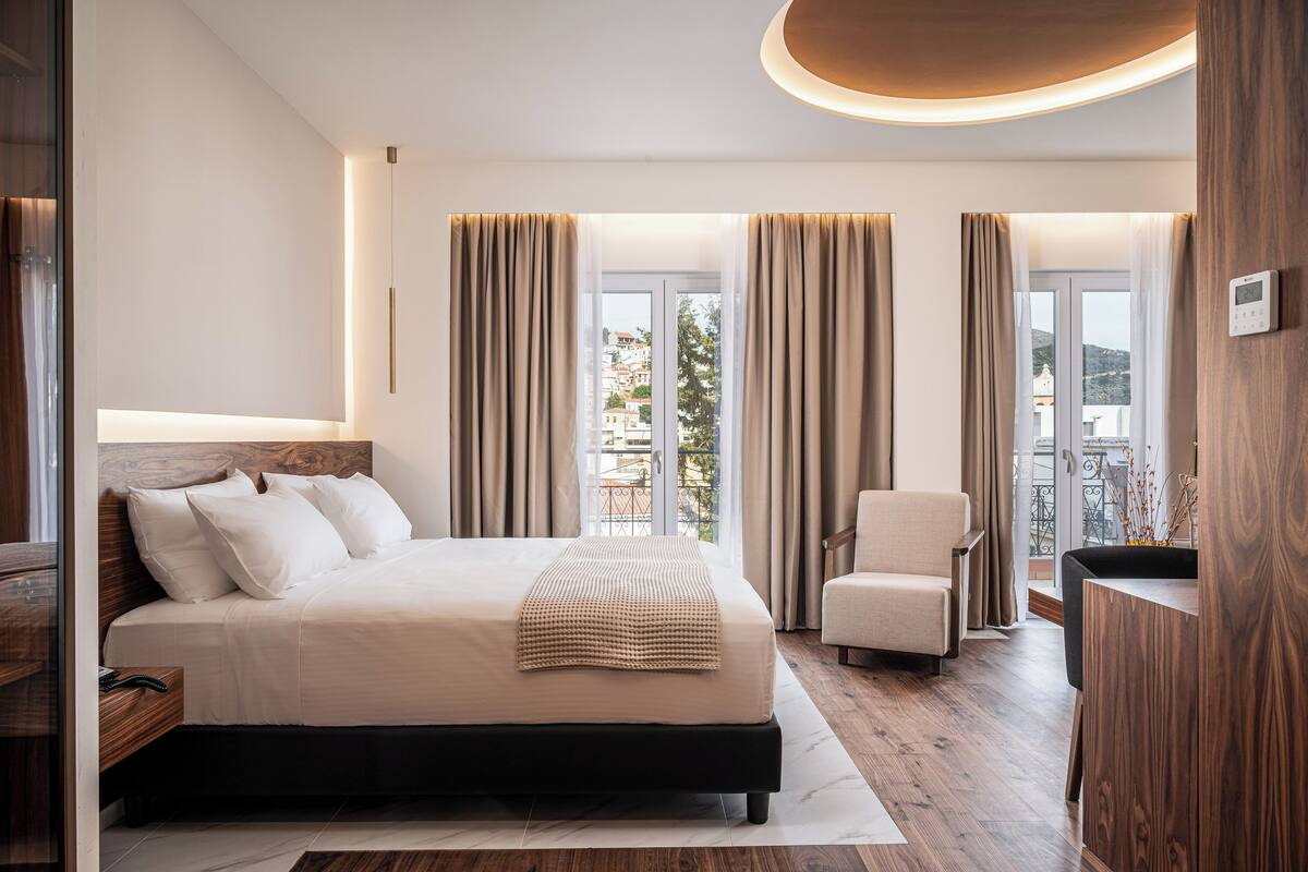 A modern hotel room with a large bed, a sitting area, and a view of the city through the windows.