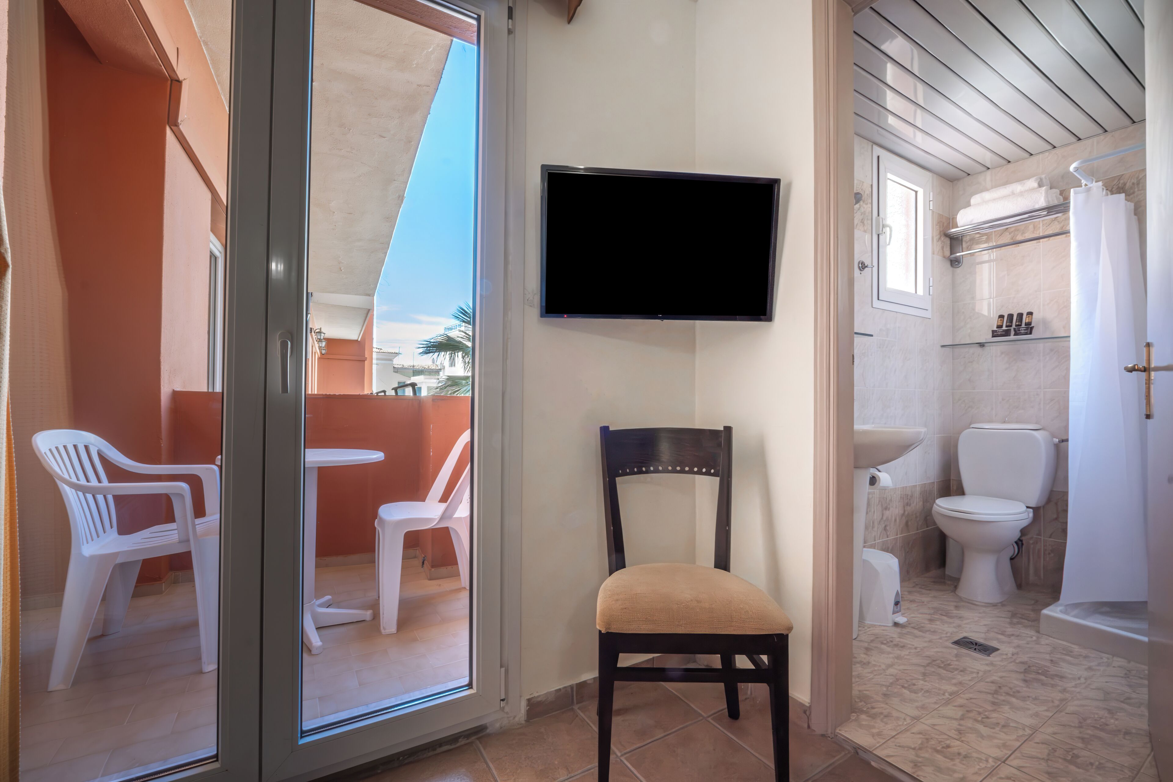 A bathroom with a toilet, sink, and shower. There is a chair and a table outside the bathroom.