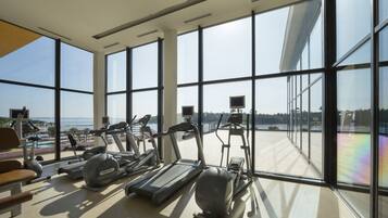 A gym with treadmills, elliptical machines, and weight benches, featuring large windows with a view of the sea.