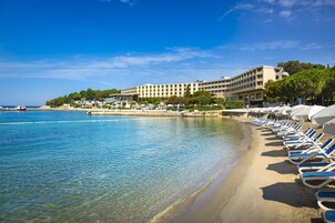 Un hotel frente a la playa con una fila de tumbonas y sombrillas a lo largo de la orilla, un mar azul cristalino y un edificio grande de varios pisos.