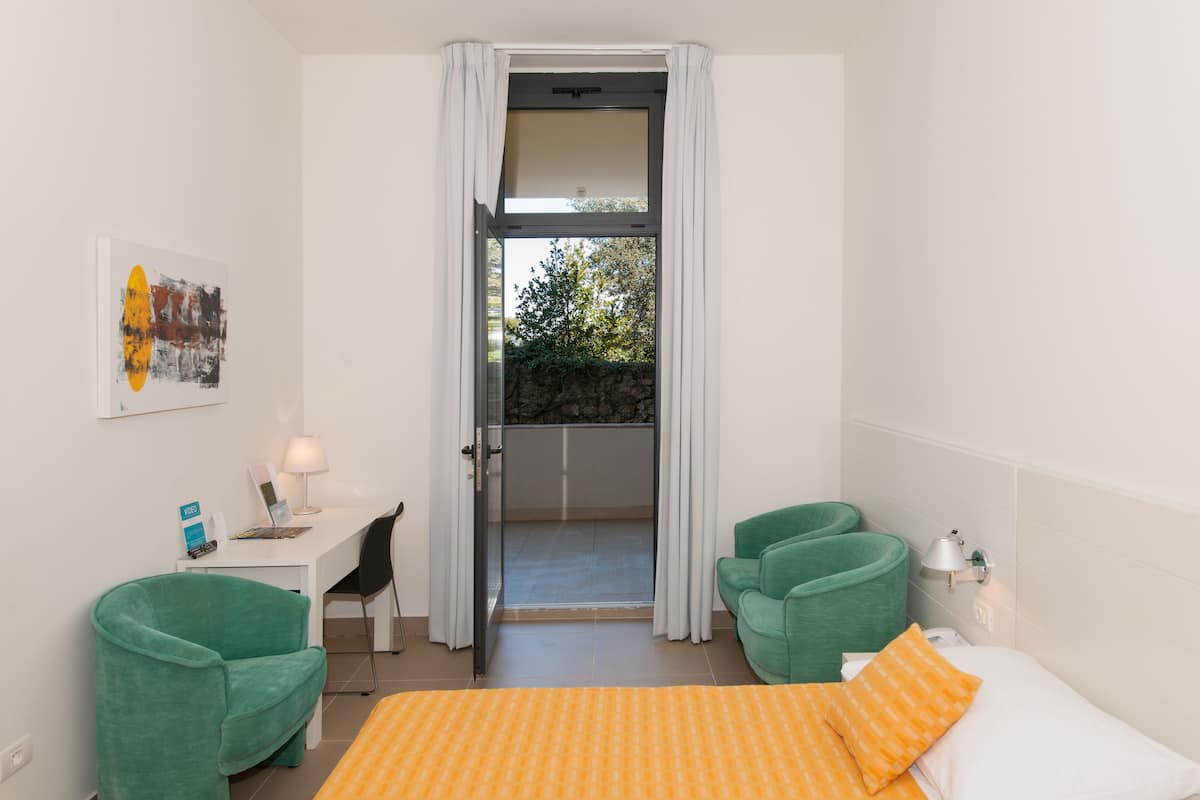 A hotel room with a bed, a desk, two green armchairs, a small table with a lamp, and a painting on the wall.