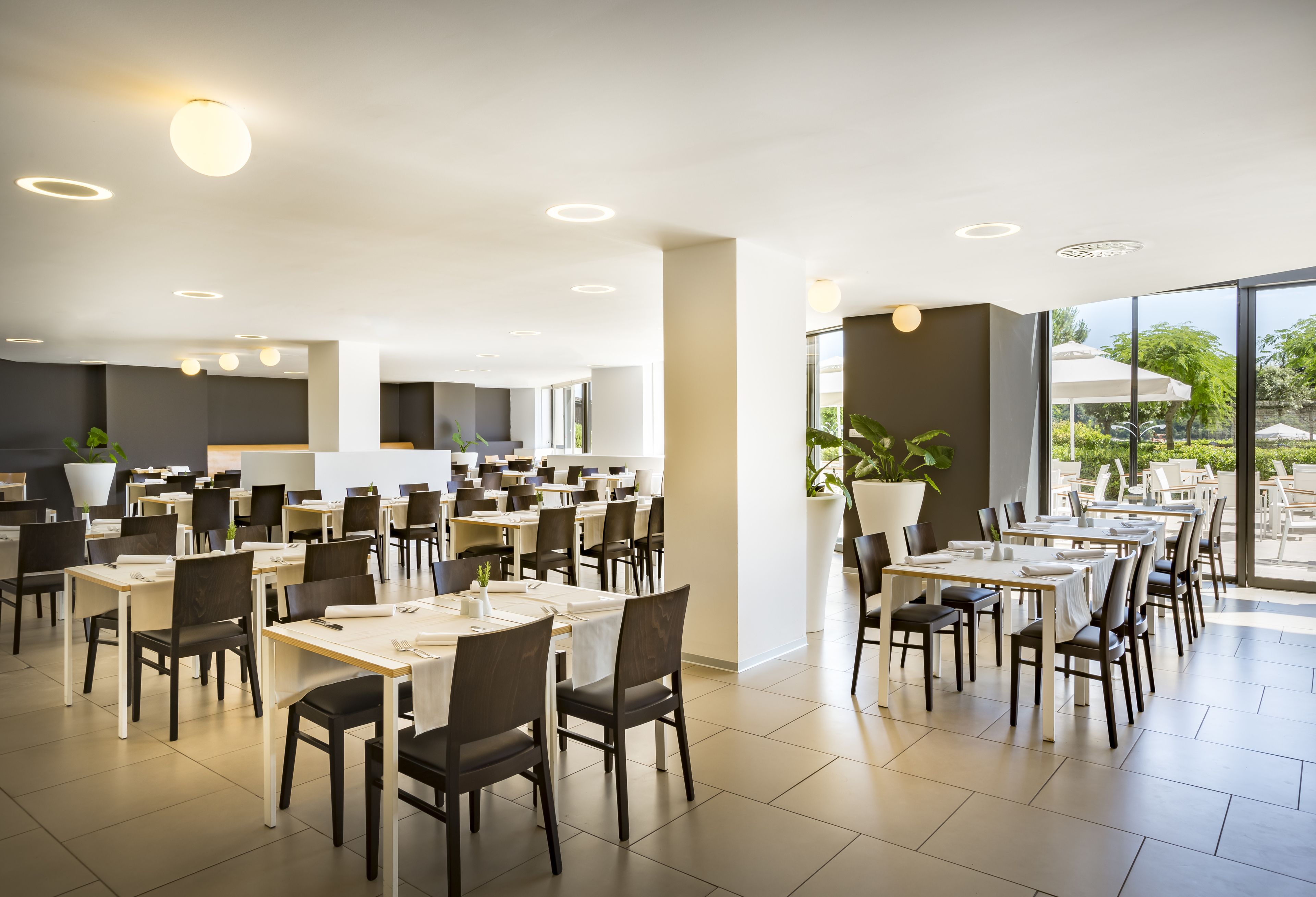 Un restaurant spacieux, doté de nombreuses tables et chaises, de grandes fenêtres et de plantes en pot.
