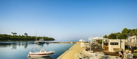 A marina with boats docked, a café area with outdoor seating, and a view of the water and greenery.