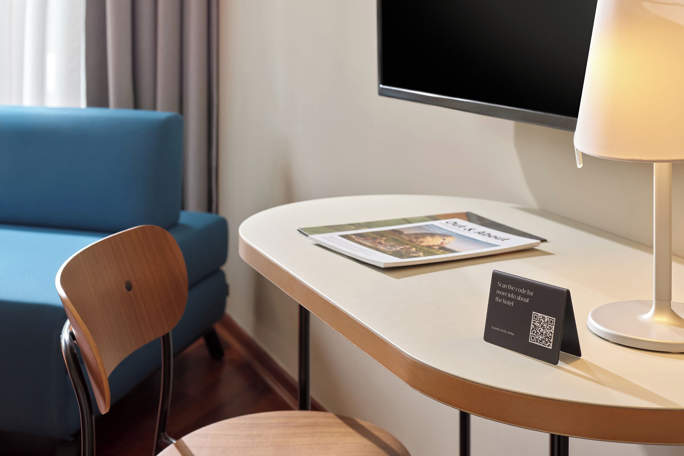 A hotel room with a flat-screen TV mounted on the wall, a small table with a lamp, a chair, and a QR code sign for hotel information.