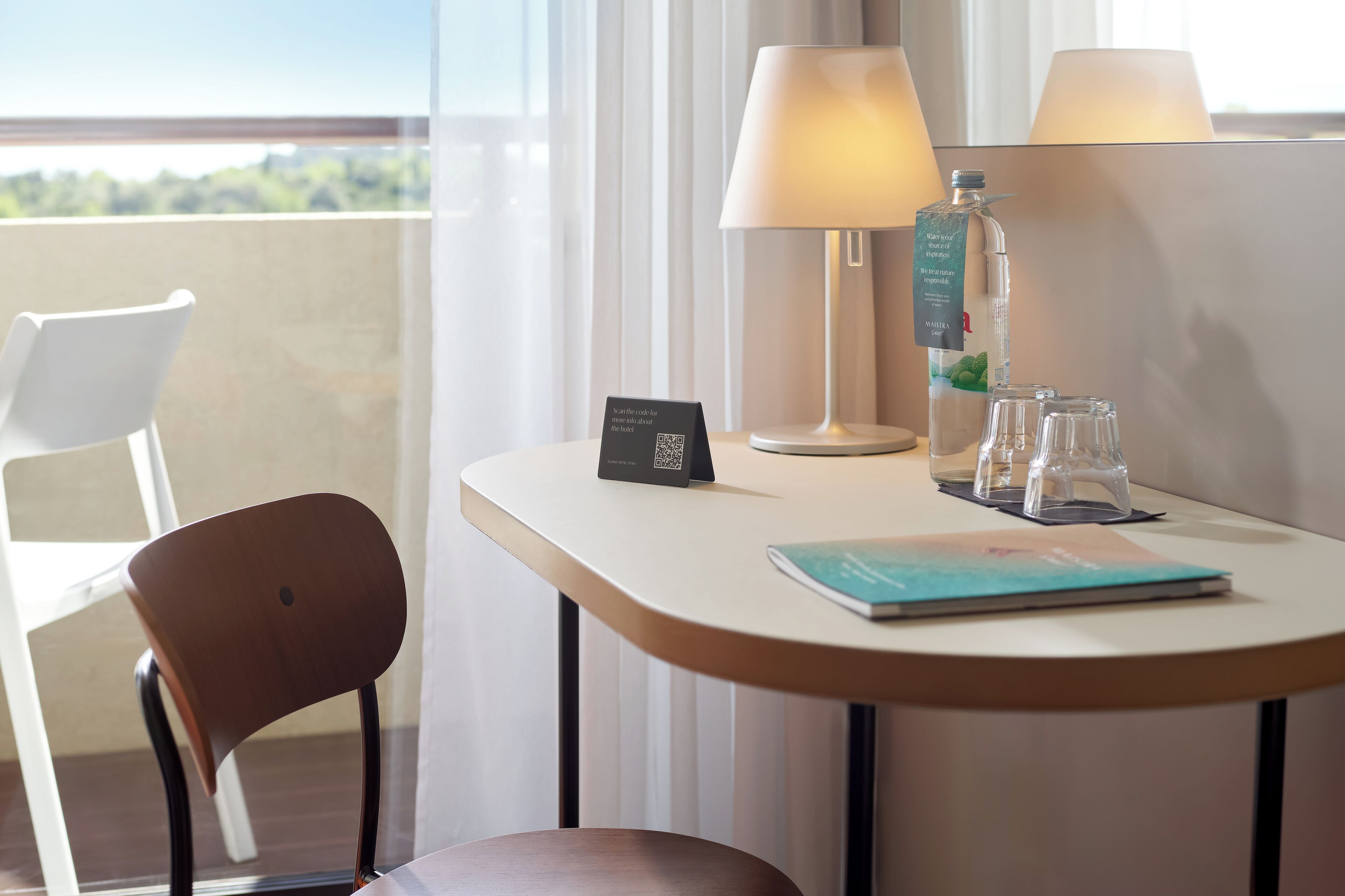 Habitación de hotel con balcón, una mesa con lámpara, una botella de agua y un cartel con un código QR.