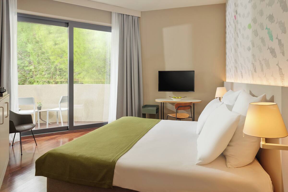 A hotel room with a bed, a TV, a desk with a chair, and a balcony with a view of trees.