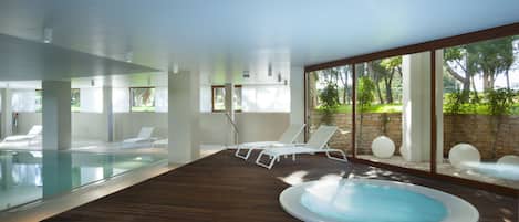 A modern indoor pool area with a hot tub, lounge chairs, and large windows offering views of greenery.