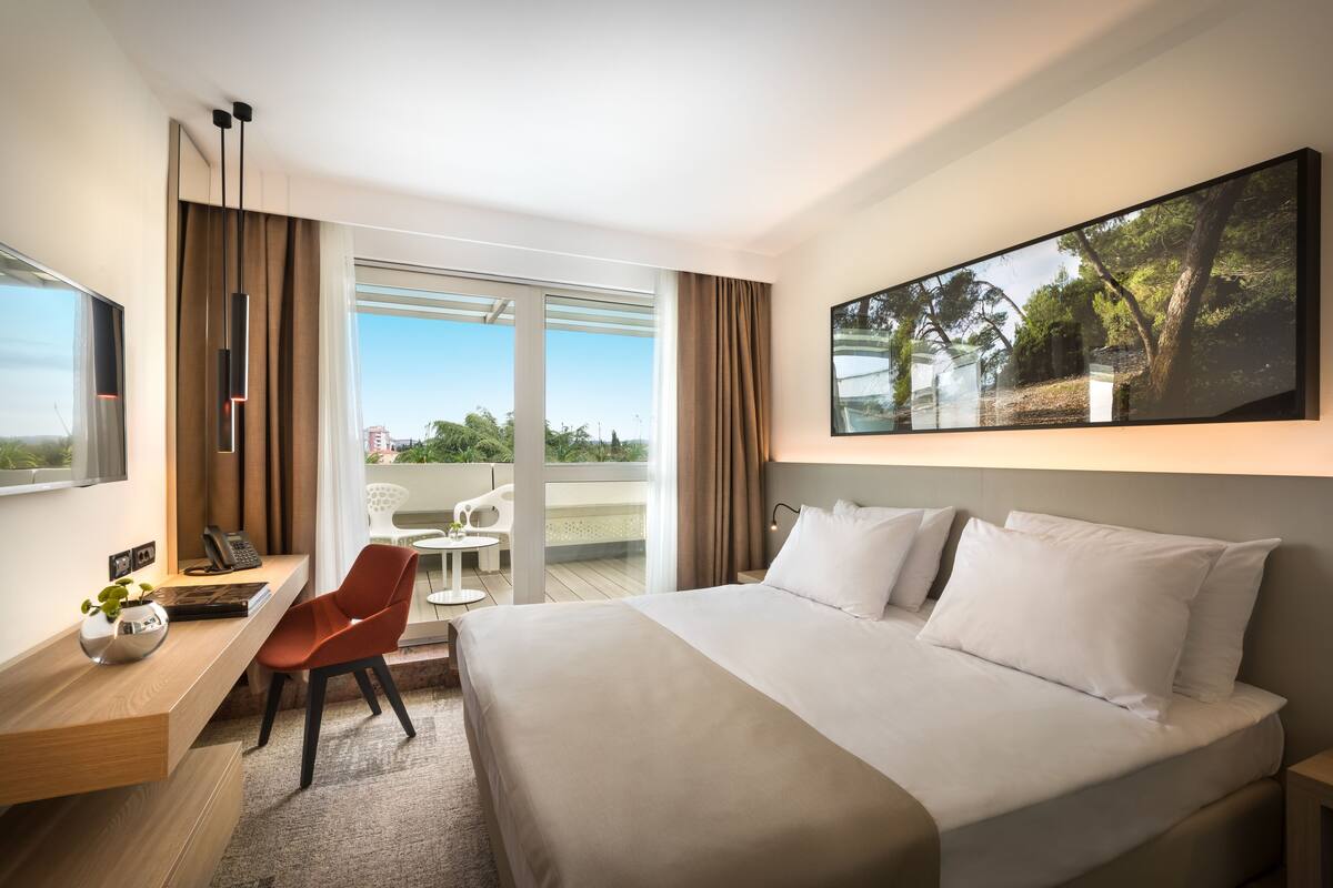 A modern hotel room with a large bed, a desk with a chair, a small table, and a large window with a view of greenery.