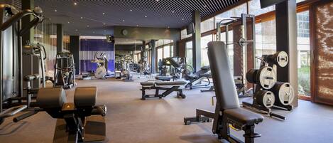 A modern gym with various exercise equipment, large windows, and a high ceiling with a grid pattern.
